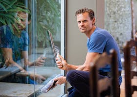 A man in a blue t-shirt cleaning large glass pane with a squeegee indoors.